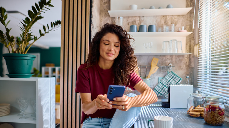 A young woman scrolls her phone while sitting at her kitchen counter