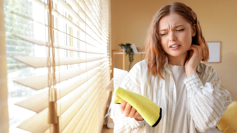 Woman about to dust blinds with a cloth, looking uncomfortable