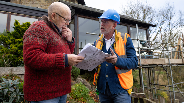 Construction worker holding blueprint and advising old homeowner with a home under renovation behind them