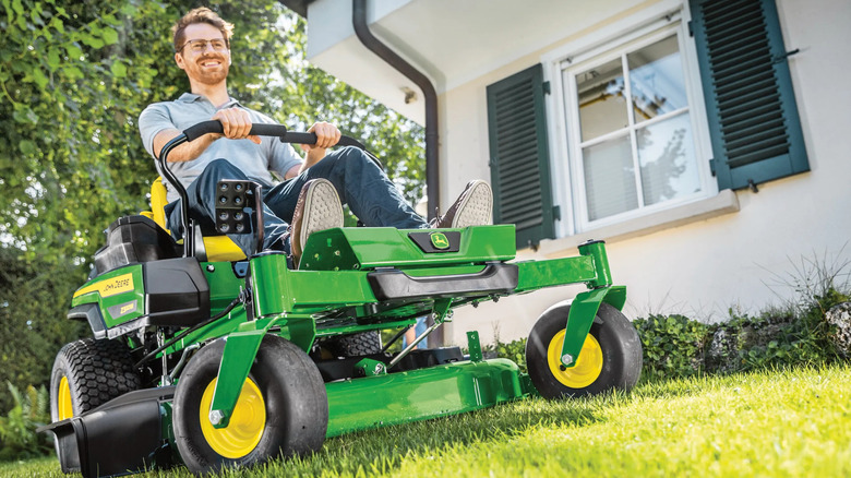 A man smiles while riding a John Deere Z320M to cut the grass in front of a white house with green shutters
