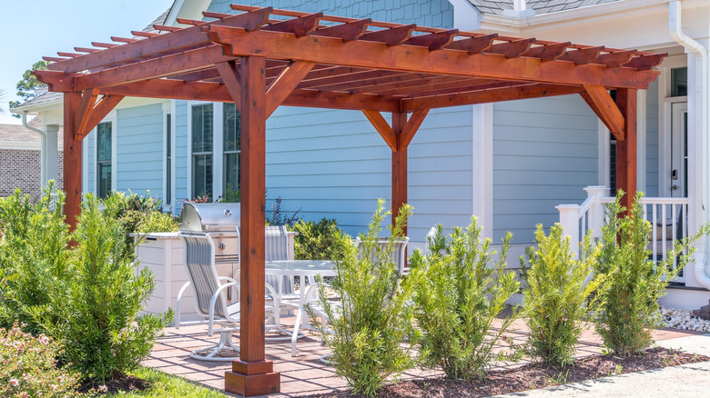 A large wooden pergola provides shade over an outdoor dining area in a suburban back yard.