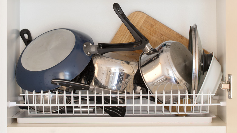 pile of cluttered pots, pans, cutting board, and lids on a cabinet drying shelf