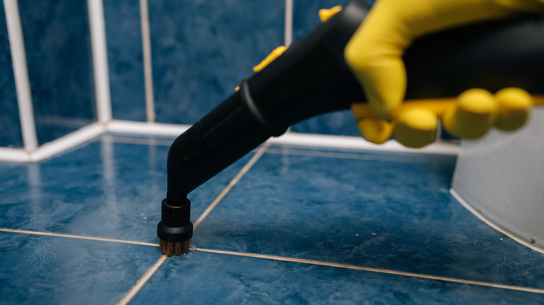 A yellow-gloved hand uses a steam cleaner on grout and blue tile