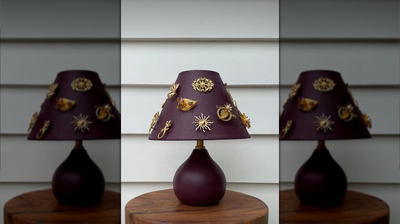 A burgundy table lamp with a collection of gold toned brooches pinned to the shade.