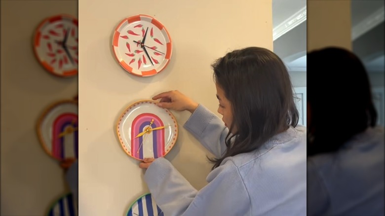 Young woman hanging gallery of handmade clocks from DIY painted plates on empty wall