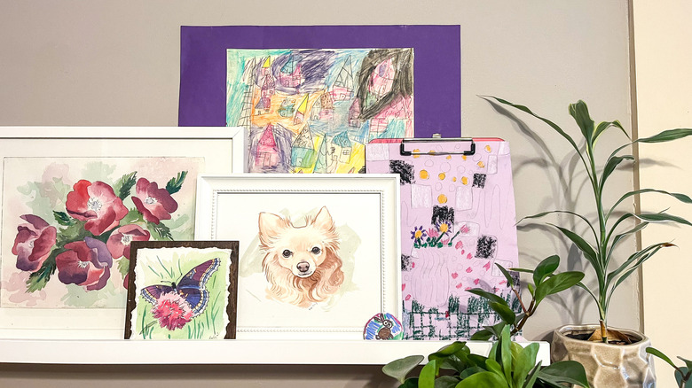 white IKEA picture ledge with an assortment of paintings and kid's artwork, one piece of which is being held up by a clipboard
