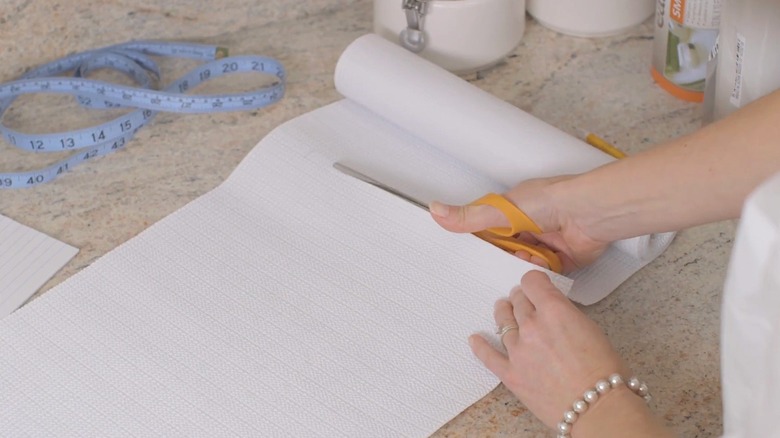 Elegant woman cutting white cushioned shelf liner with standard scissors on stone countertop