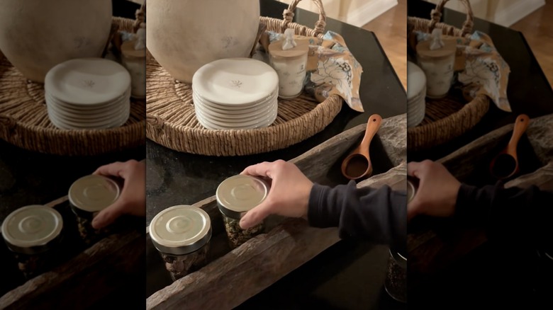 A woman's hand holding spice jars that are in a rustic dough bowl on the kitchen counter next to natural items like a jute tray and white dishes