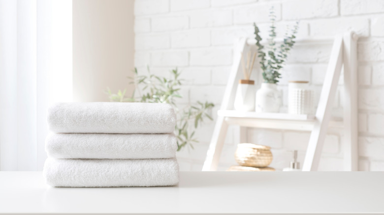 A neatly folded stack of white towels on a bathroom counter, with white tile and shelf in background.