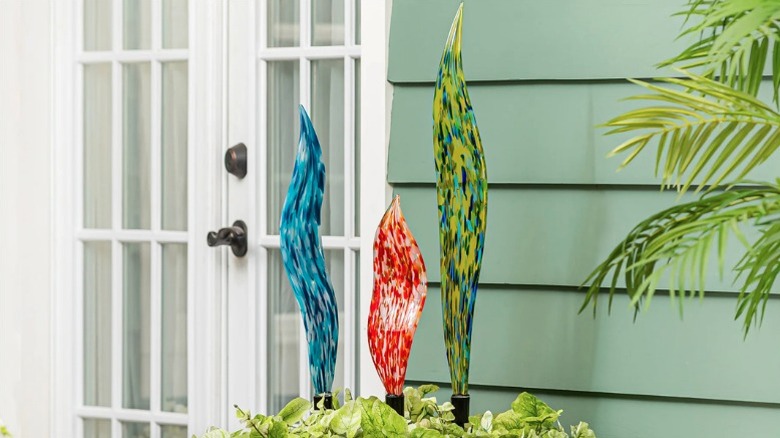 Three colorful hand blown glass garden stakes decorate a terracotta planter in front of a sage green house