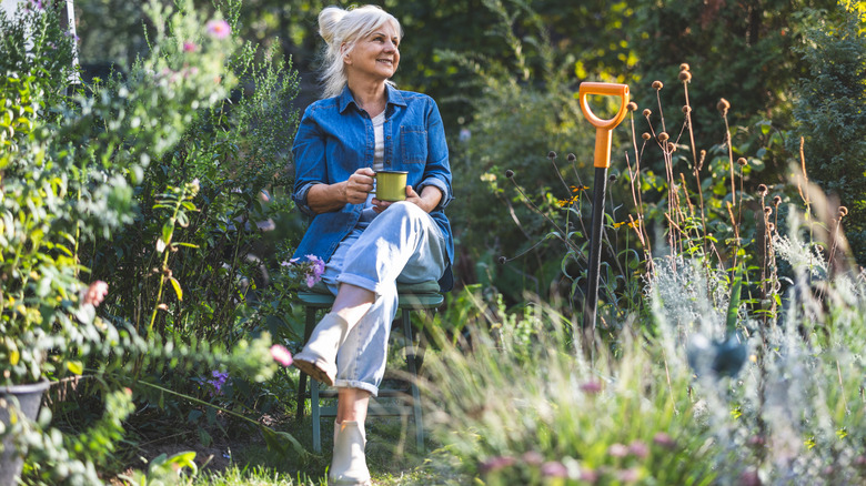 An older woman holds a mug while sitting on a bench in the garden