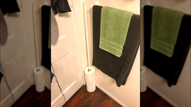 Toilet paper being stored on a broom handle in the corner of a bathroom