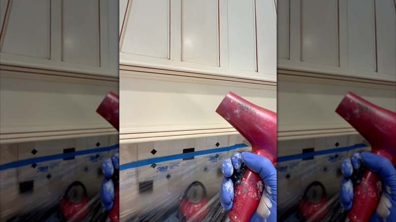 Someone wearing a blue plastic glove and pointing a red hair dryer pointed at kitchen cabinet