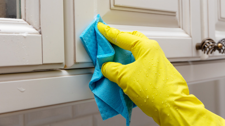 Someone's hand wearing a rubber glove and scrubbing a greasy kitchen cabinet with a blue cloth