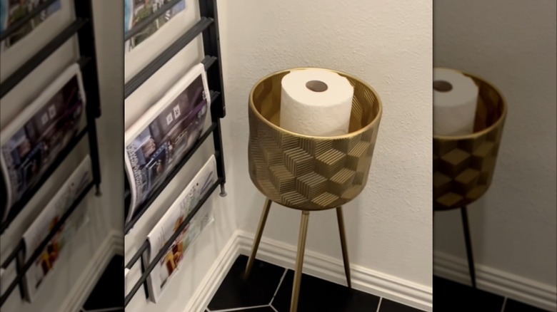 A gold plant stand with a roll of toilet paper next to a magazine rack on the wall and on a black tiled floor