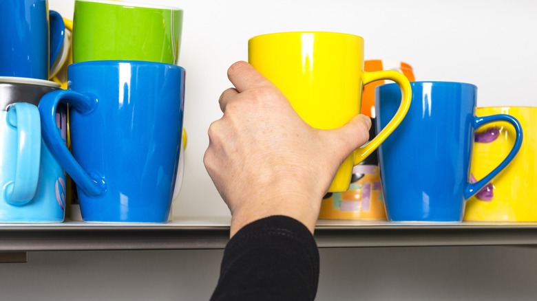 Person grabbing yellow mug from kitchen shelf full of colorful coffee mugs