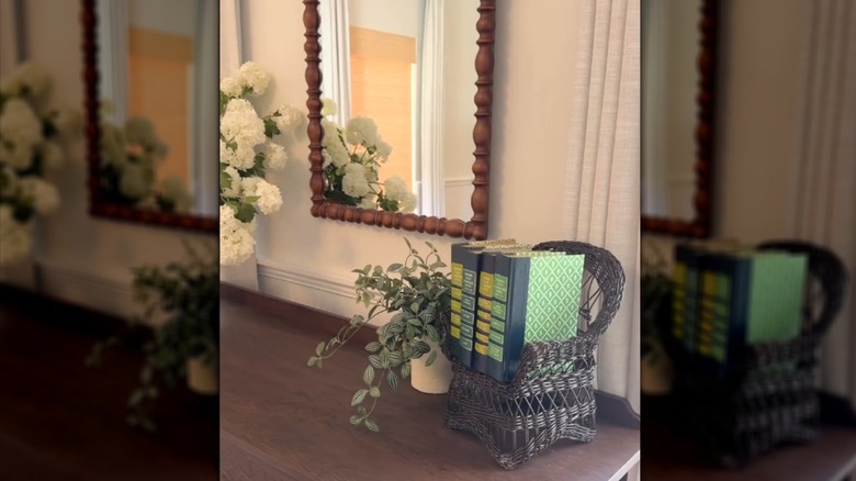 Thrifted brown wicker doll chair with small books on solid wood dresser top with plants and wood framed mirror
