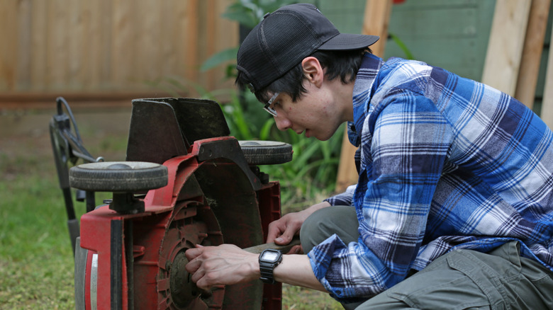 A person wearing a flannel shirt removes blades from a push lawnmower