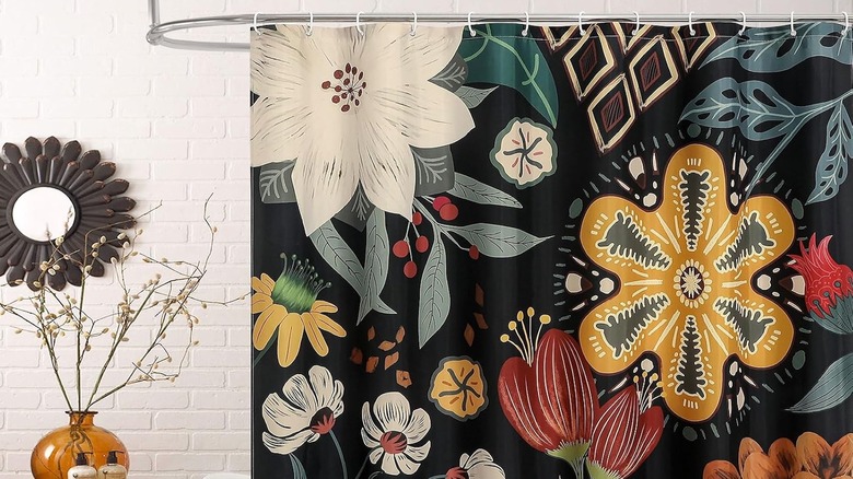 A black floral shower curtain in a boho bathroom with a white brick wall, statement mirror, and nearby wooden table
