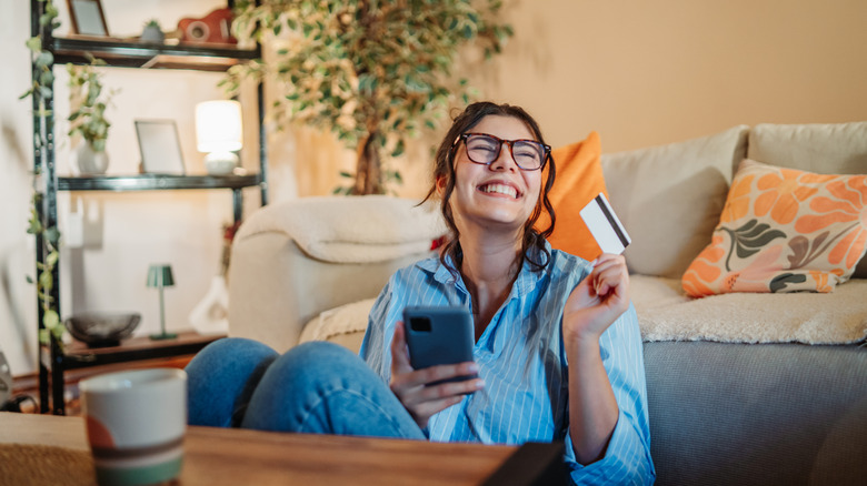 A woman smiles while shopping on her phone, with a credit card in one hand