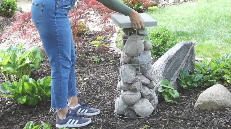 Woman in cropped jeans piling decorative rocks in wire tomato cage in landscaped yard