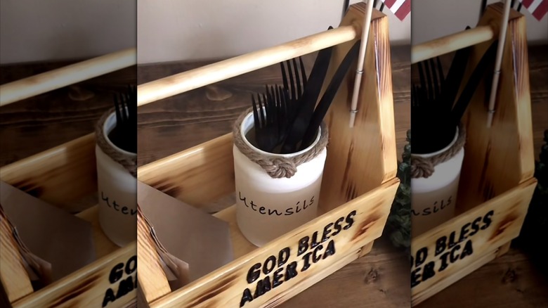 A white DIY utensil holder in a wooden tray with the word "utensils" on it and plastic, utensils in place