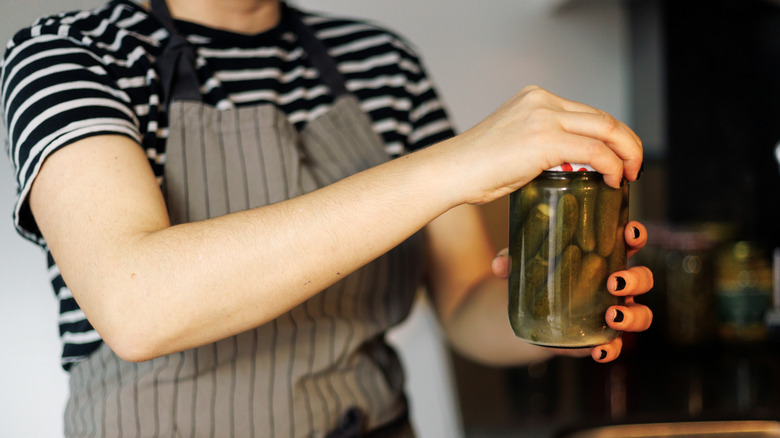 A faceless woman in a black and white striped shirt and apron in the kitchen opening a pickle jar