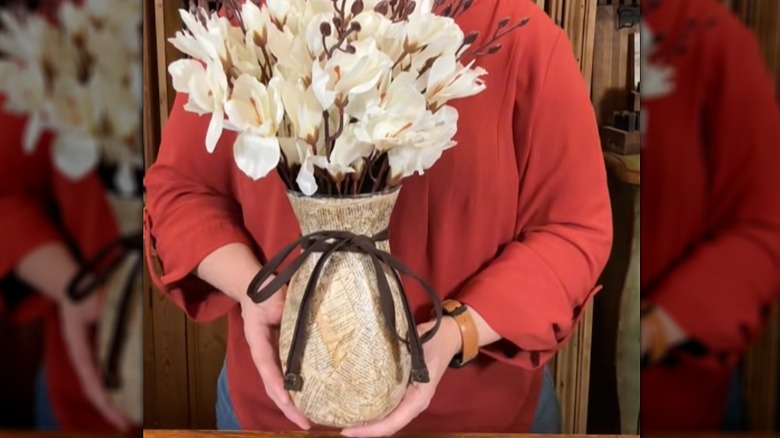 Creator holding a glass vase covered in book pages with a ribbon tied around it and white flowers inside