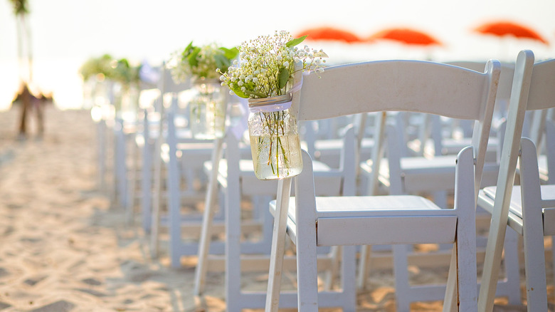 mason jars with flowers hung from white chairs at a ceremony on the beach