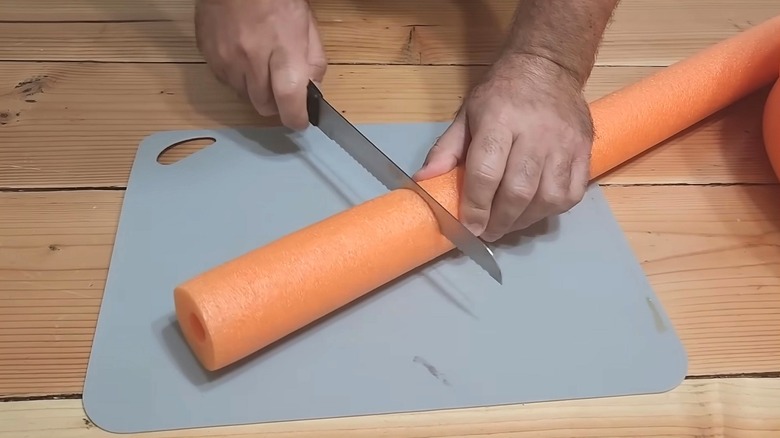 A person slices an orange pool noodle with a bread knife on a gray cutting board