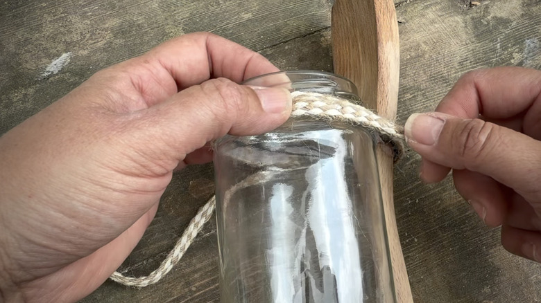 A woman wrapping jute cord around a glass jar and wooden spoon on a distressed wooden table