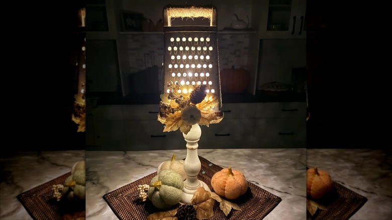 A DIY lamp made from an old cheese grater and empty pickle jar sits on a counter surrounded by fall decor pumpkins
