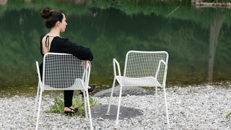 A dark haired woman sits in a white metal chair overlooking a lake