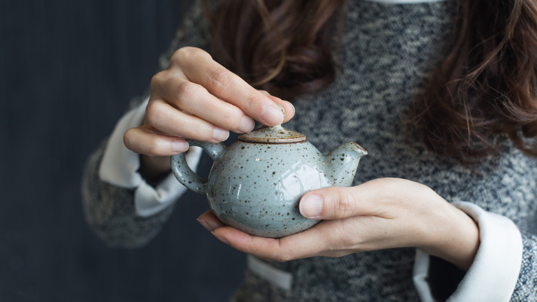 person wearing a grey sweater and holding an old tea kettle with both hands