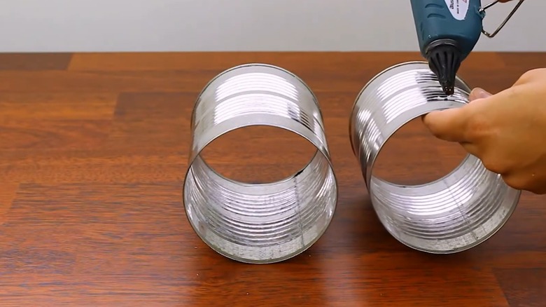 Woman hot-gluing two hollowed-out metal soup cans together on dark red wood table