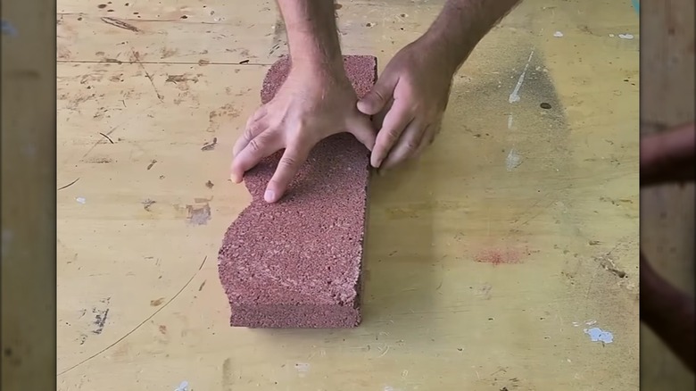 Man holding red scalloped concrete edging stone against dirty wood work surface