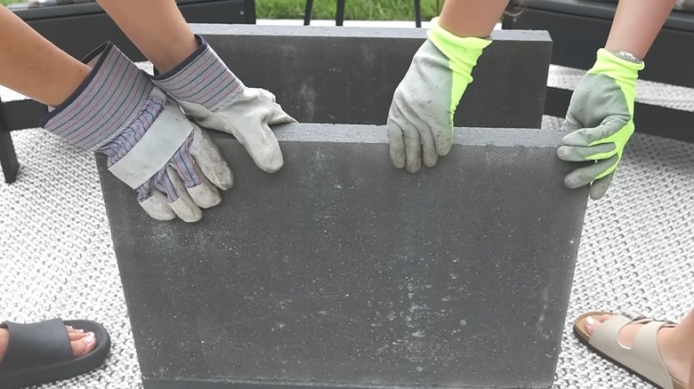 Couple lifting heavy paver with gloves on outdoor deck with carpet