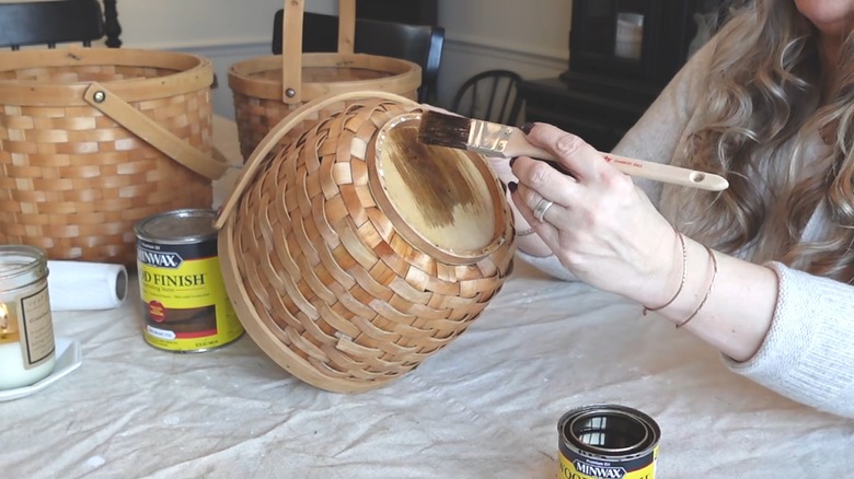 Woman staining woven wicker baskets with dark wood stain over tarp on table