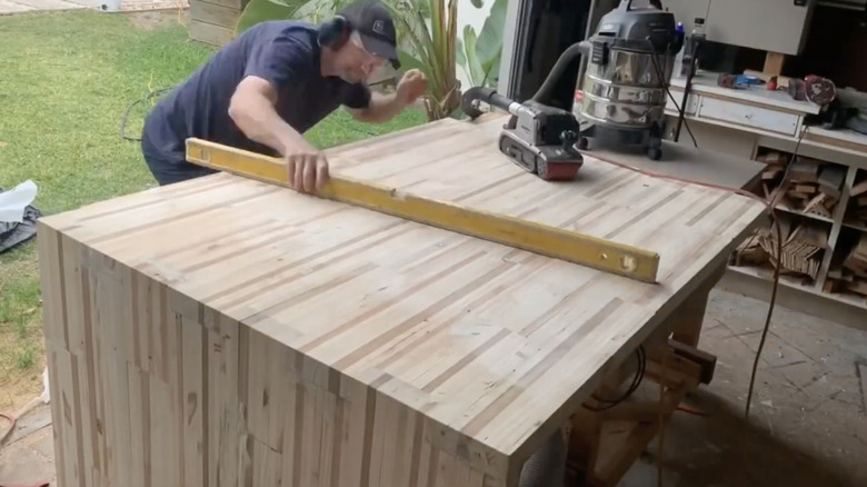 Man checking pallet wood island top for level in garage shop with backyard in view.