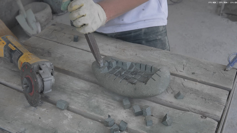 A person wearing gloves chiseling out a depression in river rock with squares cut into it with an angle grinder