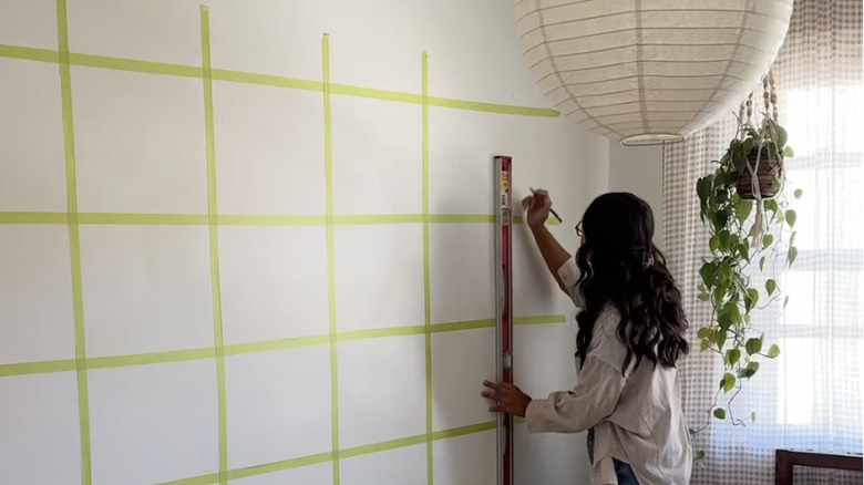 DIYer using a level, painter's tape, and a pencil to mark out a grid on the wall for building a floating shelving unit