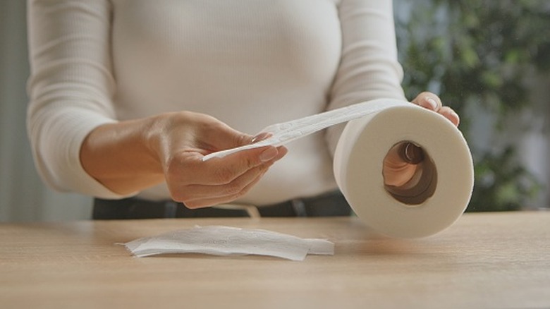 A woman sits at a table while holding a roll of toilet paper