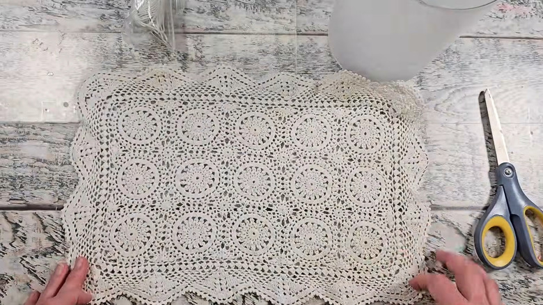 A DIYer prepares a lace doily on a wood table, with a pair of scissors nearby