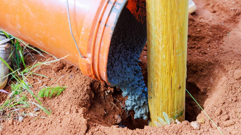 Person pouring concrete from a bucket into a hole with a post
