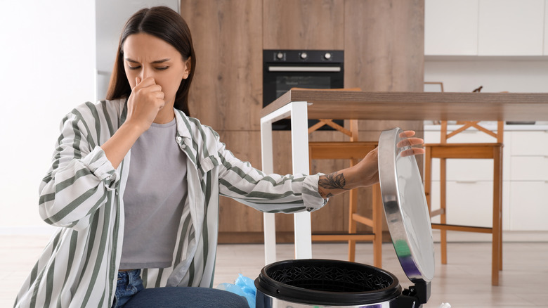 A woman holds her nose as she opens a garbage can.