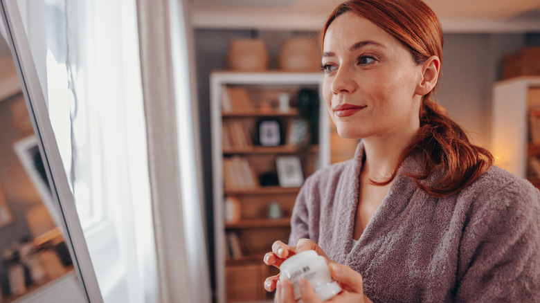 A woman in a bathrobe holding a skincare jar.