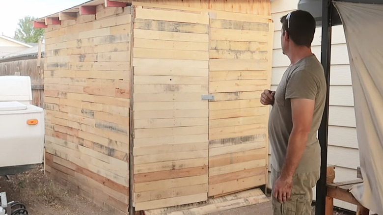A man standing next to a storage shed built out of wood pallets