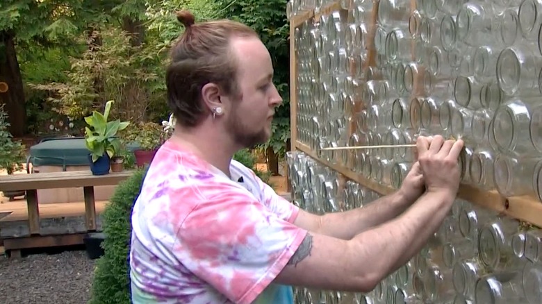 A person wearing a tie-dye shirt uses a stick to poke moss into a greenhouse made of upcycled glass jars
