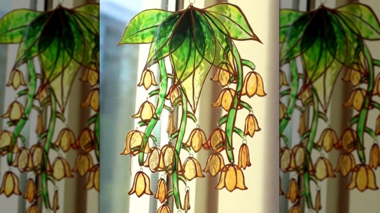 Sun catcher with green leaves, vines, and bell-like gold flowers made from plastic cups hangs in window.