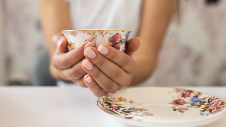 A person with a pink manicure holding a vintage floral teacup over a saucer